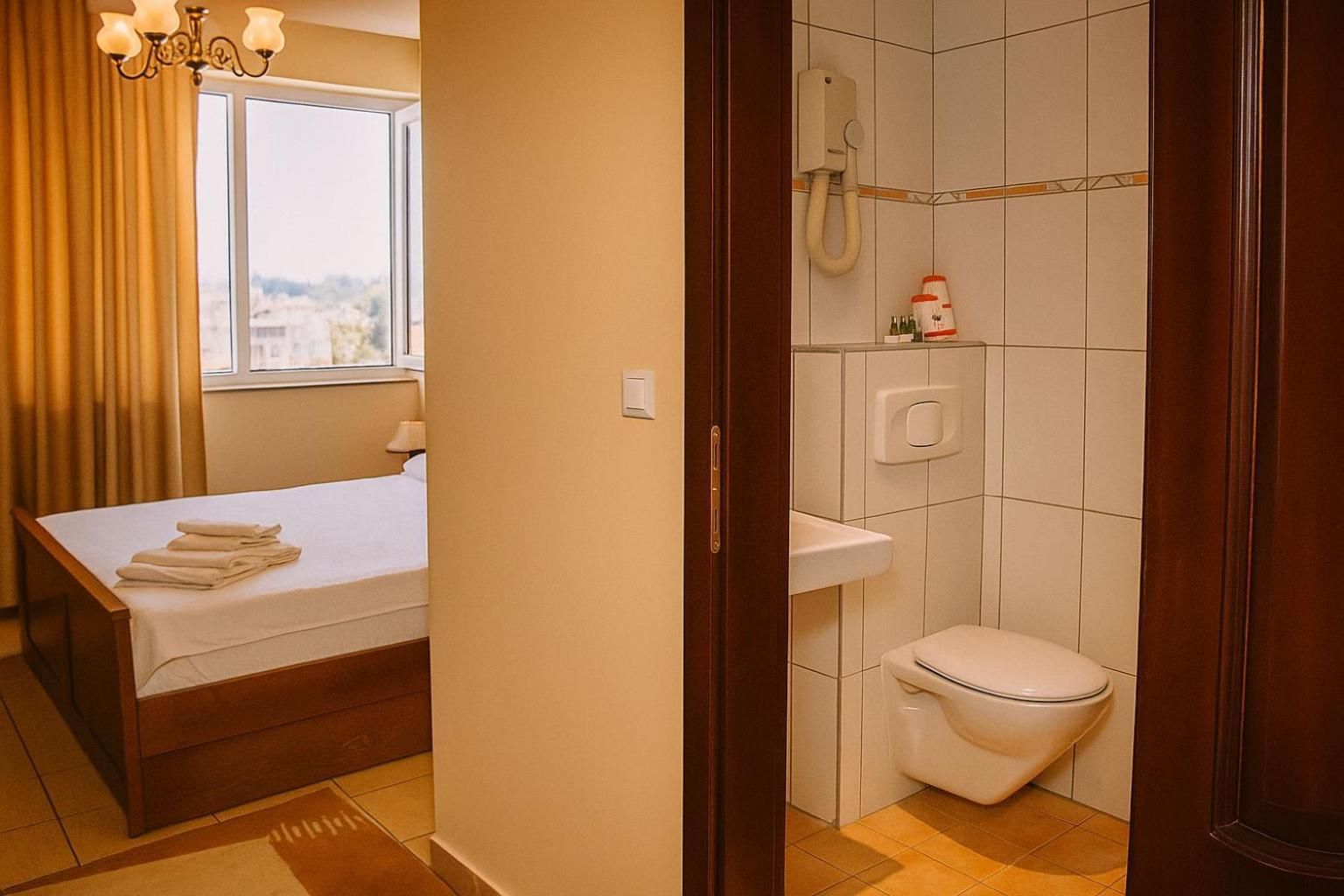 A hotel room with a bed, a window with curtains, a shower area with a toilet, and a towel rack.