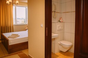 A hotel room with a bed, a window with curtains, a shower area with a toilet, and a towel rack.