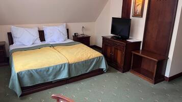 Una cama doble con colcha verde y amarilla, un televisor sobre un mueble de madera, y un armario de madera con ganchos.