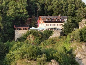 A multi-story building with a stone wall and a red roof, surrounded by dense forest. - Naturfreundehaus Veilbronn (Heiligenstadt in Oberfranken)