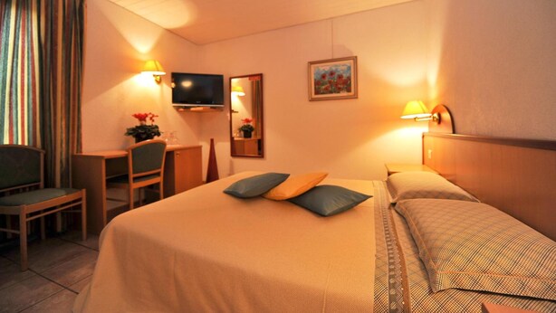 A hotel room with a bed, a desk, a chair, a television, and a painting on the wall. - Hotel Ristorante Grotto Serta (Lamone)