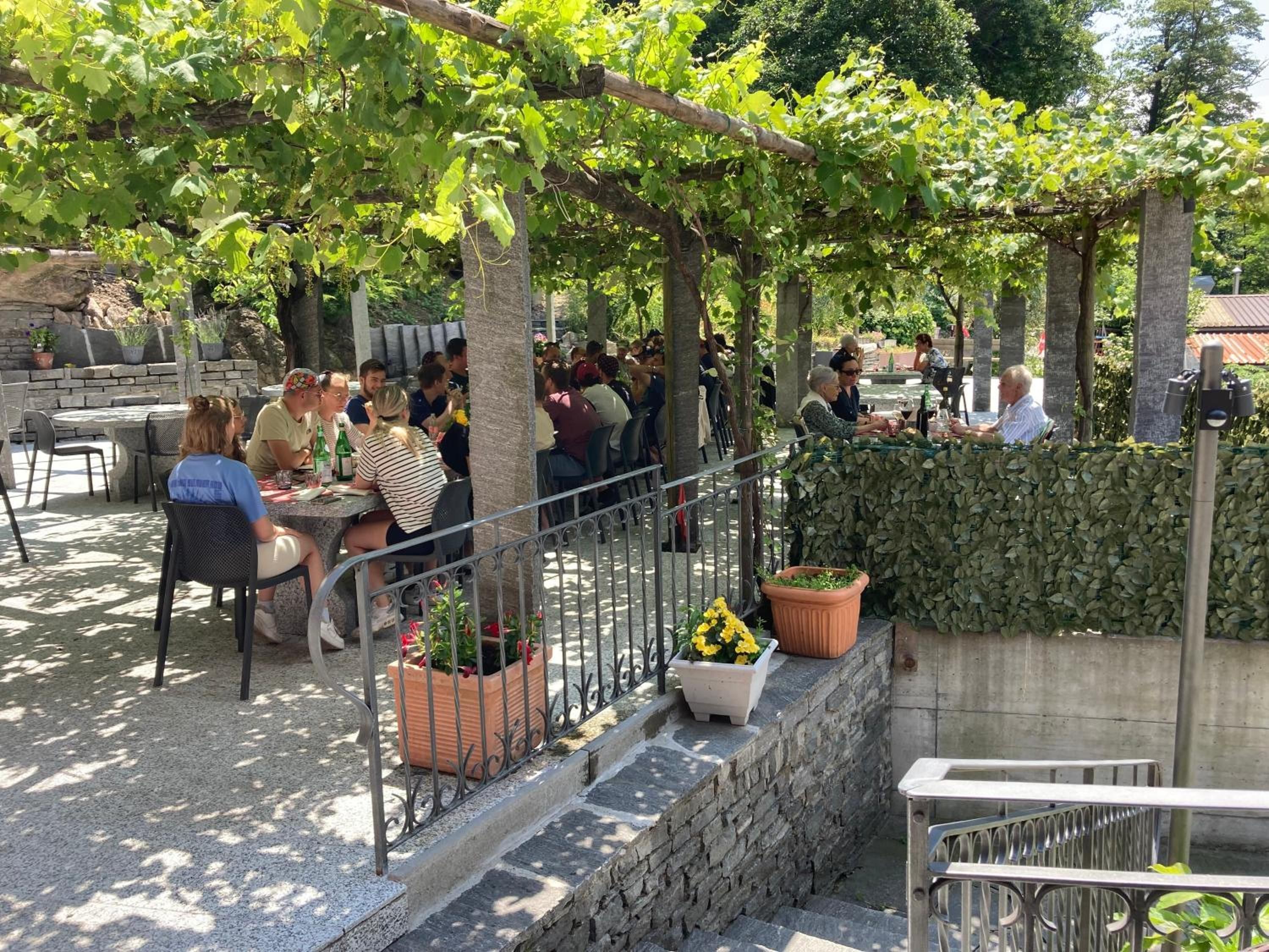 An outdoor seating area with tables and chairs, surrounded by greenery and stone walls.