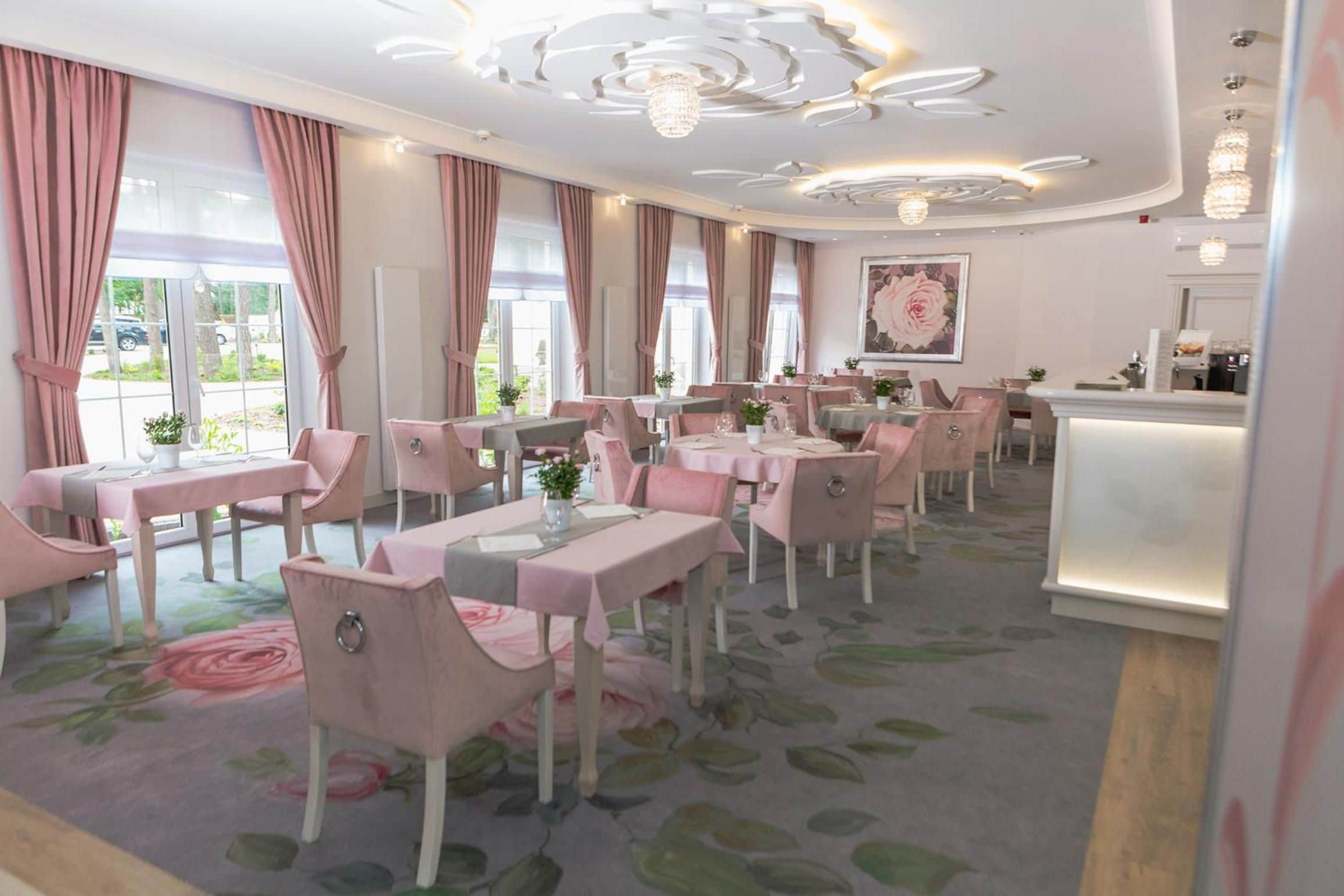 En restaurant med rosa indretning, herunder stole, borde og gardiner. Der er flere spiseborde med stole, en lysekrone og et stort vindue med gardiner.