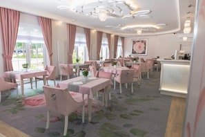 A restaurant with pink-themed decor, including chairs, tables, and curtains. There are multiple dining tables with chairs, a chandelier, and a large window with curtains.