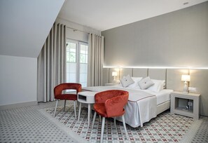 Habitación de hotel moderna con una cama, dos sillas rojas, una mesita y una mesita de noche con lámparas.