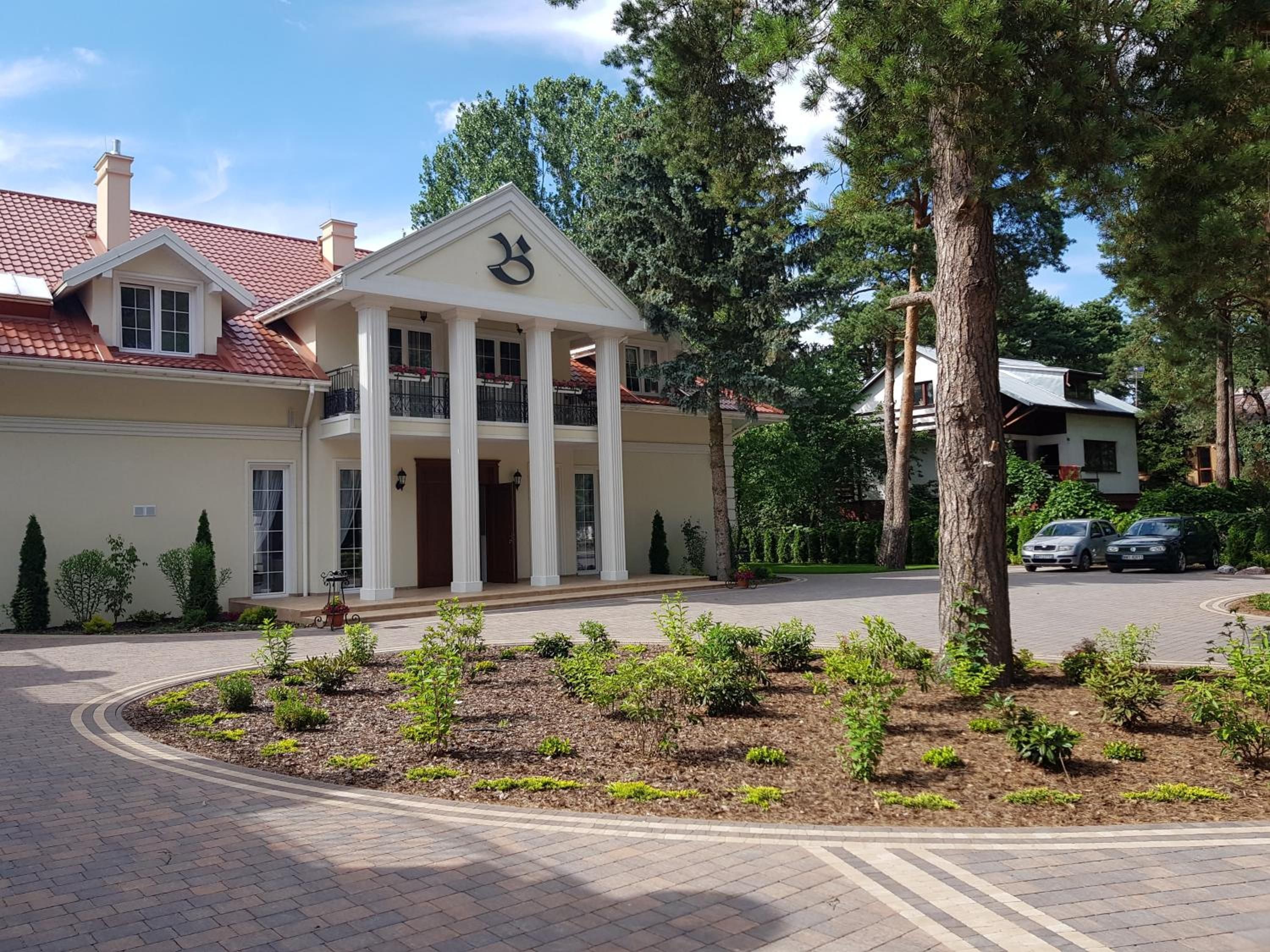 A two-story building with a central entrance flanked by columns, surrounded by a landscaped garden and a paved driveway.