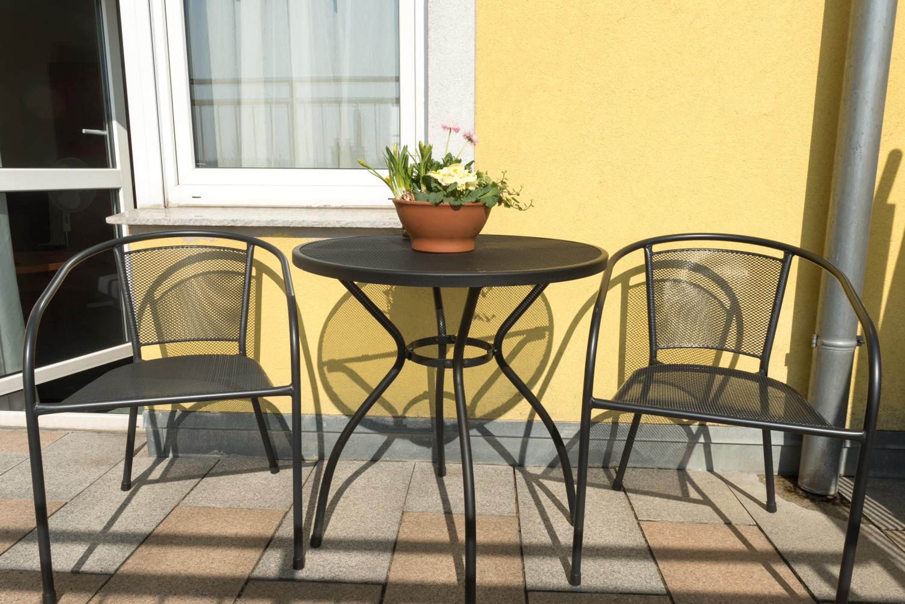 A small outdoor seating area with a round table and two chairs, a potted plant on the table, and a yellow wall in the background.