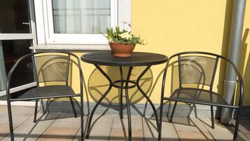 A small outdoor seating area with a round table and two chairs, a potted plant on the table, and a yellow wall in the background.