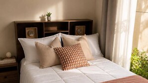 Cama bem arrumada com manta listrada, cabeceira com alto-falantes embutidos e mesa de cabeceira com abajur e um objeto decorativo.