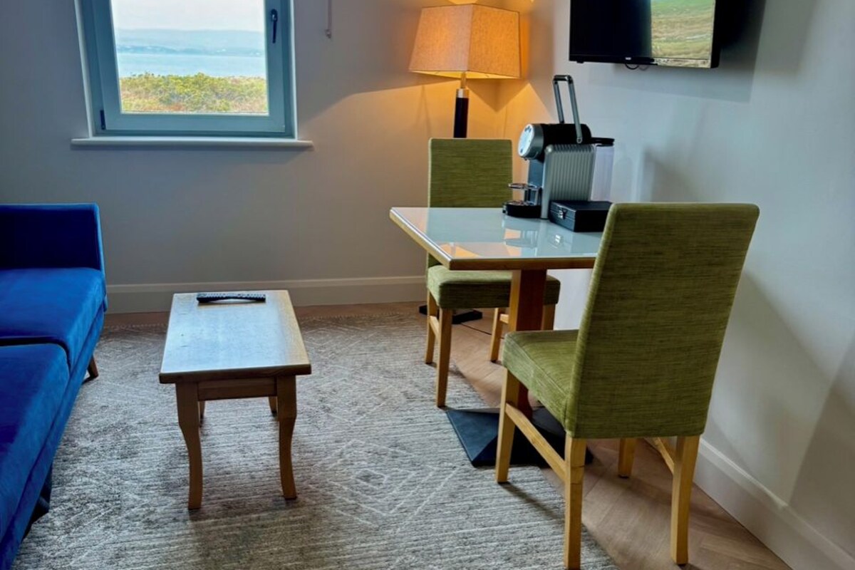 A room with a sofa, a small table, a dining table with chairs, a TV, and a window with a view.