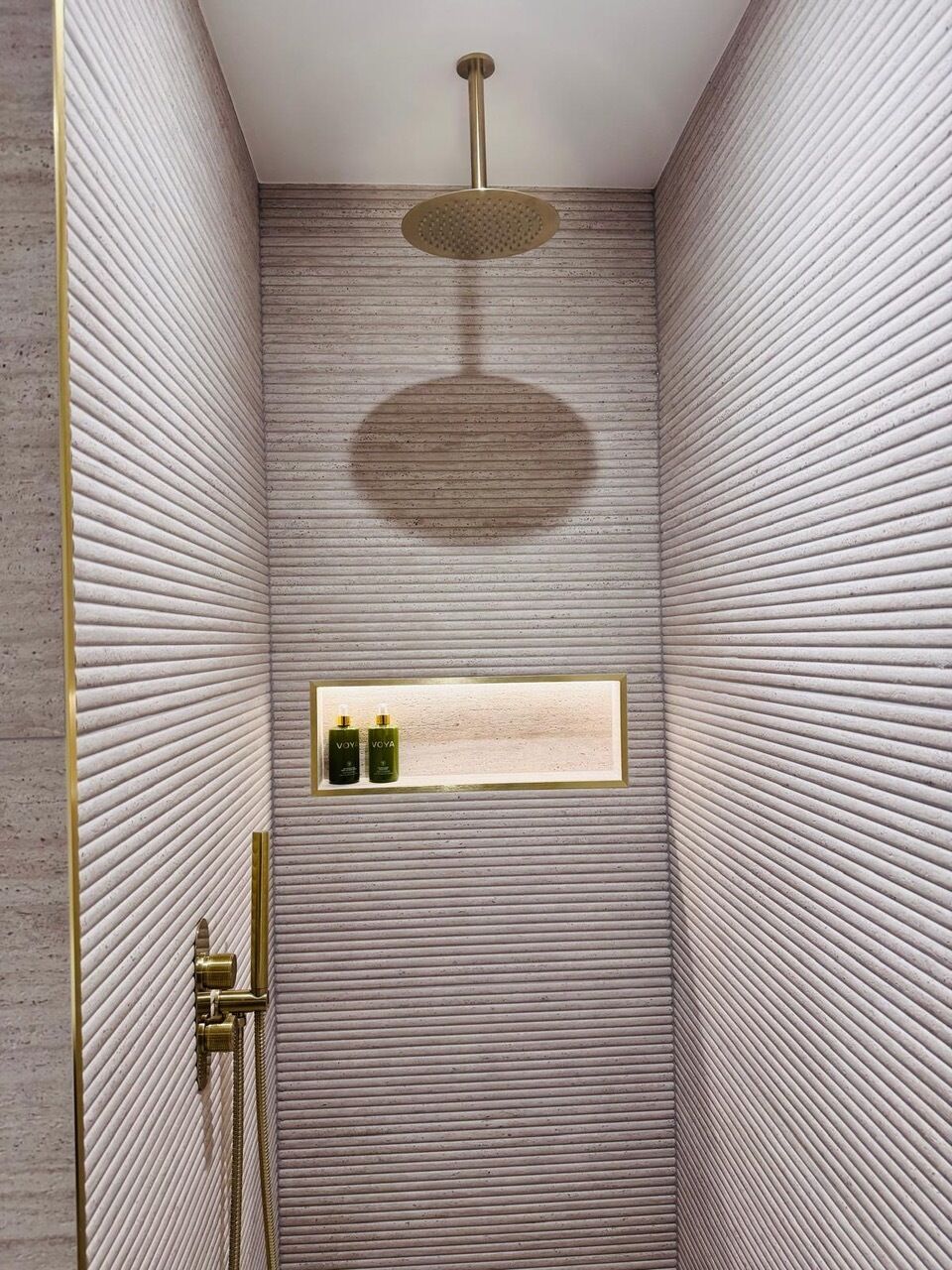 A shower with a textured wall, a gold showerhead, and a shelf with bottles.