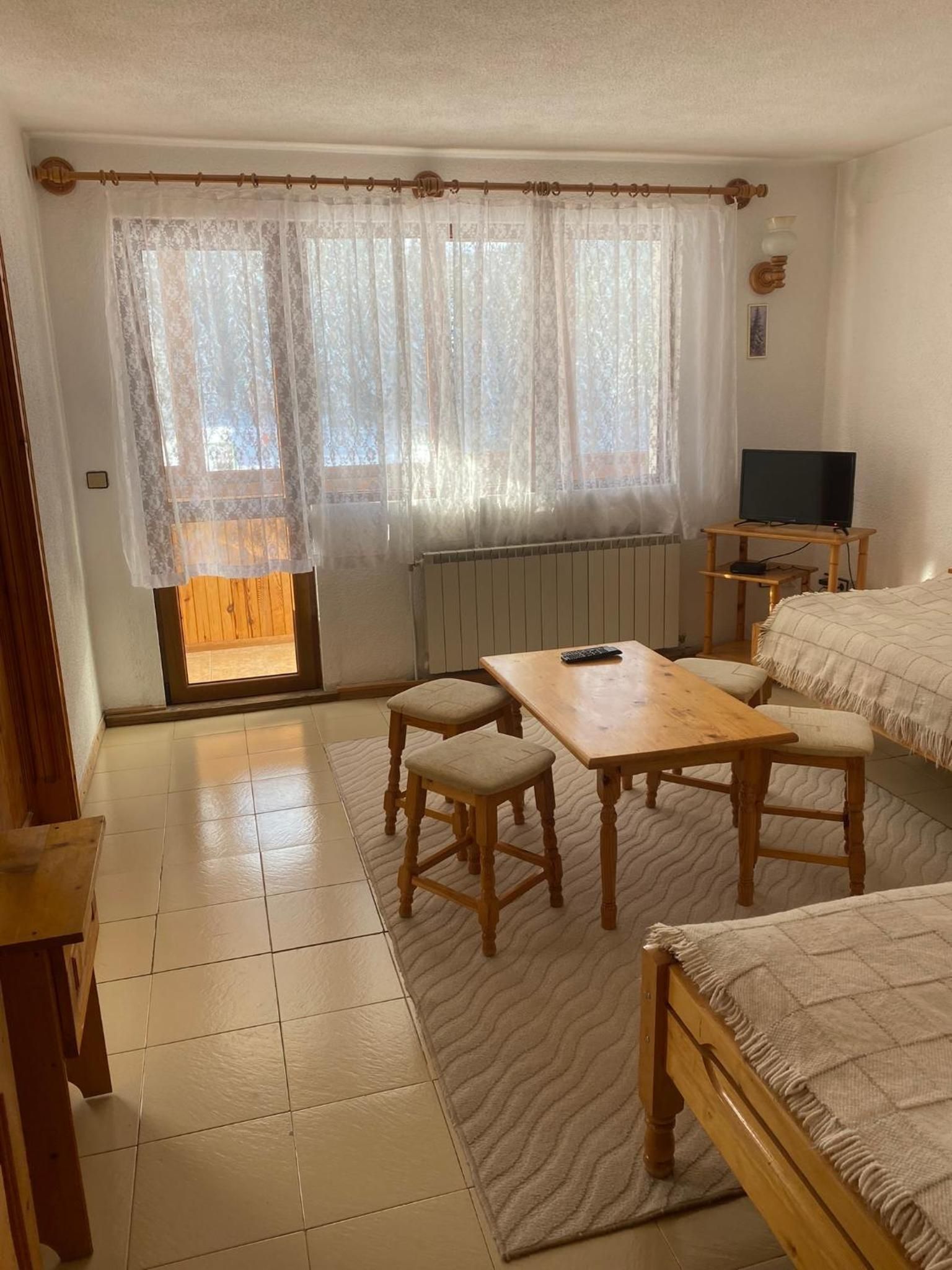 Una habitación con una mesa de madera, dos sillas, una cama, un televisor y una ventana con cortinas translúcidas.