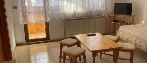 Una habitación con una mesa de madera, dos sillas, una cama, un televisor y una ventana con cortinas translúcidas.