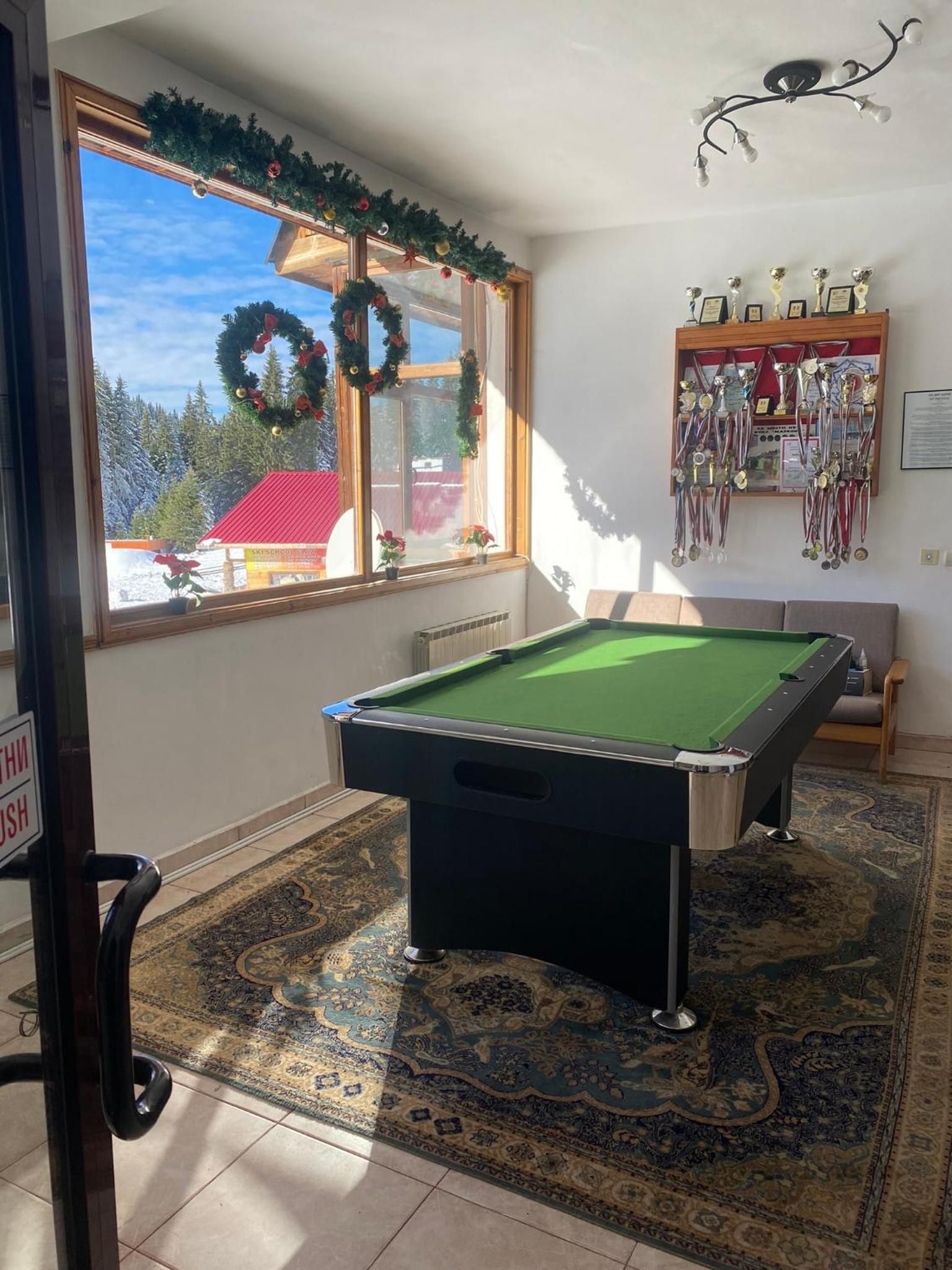 Une table de billard dans une pièce donnant sur un bâtiment, une fenêtre décorée d’une guirlande et un mur orné de trophées.