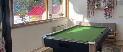 Une table de billard dans une pièce donnant sur un bâtiment, une fenêtre décorée d’une guirlande et un mur orné de trophées.