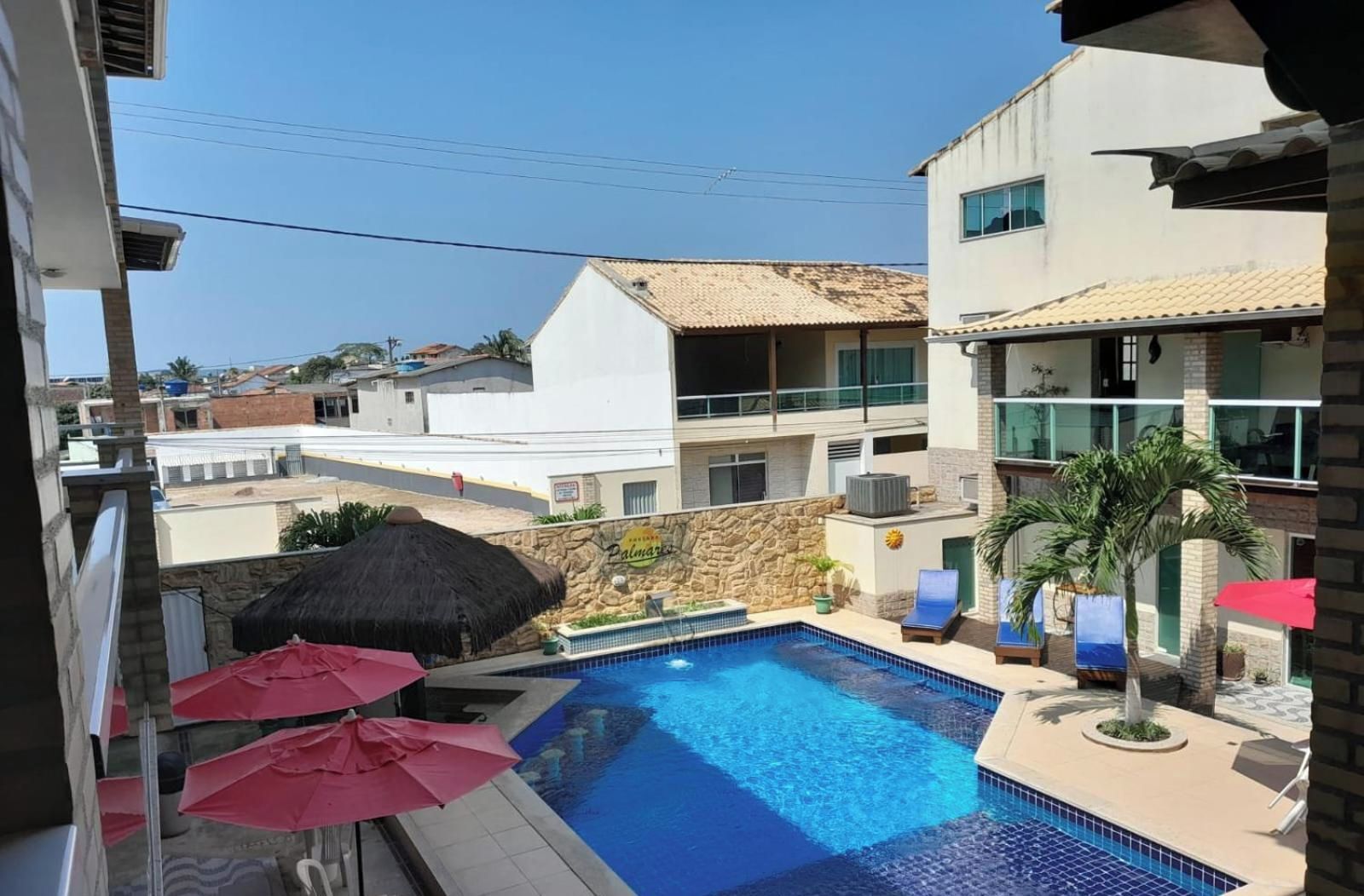 A swimming pool with lounge chairs and umbrellas, surrounded by buildings with balconies and a stone wall.