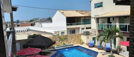 A swimming pool with lounge chairs and umbrellas, surrounded by buildings with balconies and a stone wall.