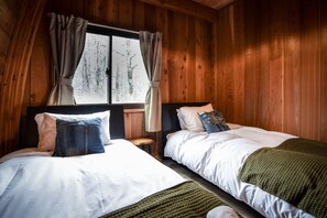 Una habitación con dos camas, paredes de madera, una ventana con cortinas y vistas a árboles.