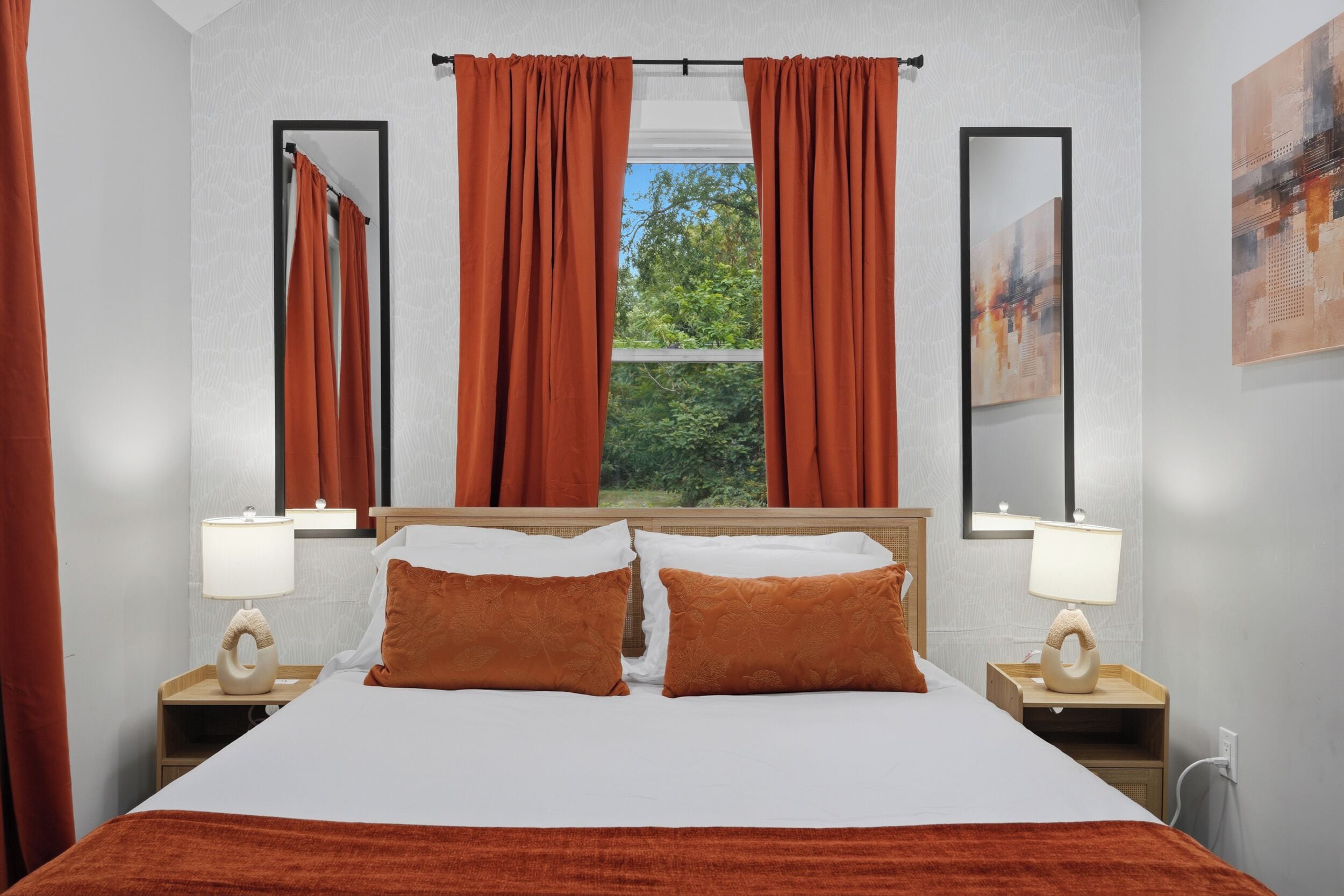 A hotel room with a bed, two pillows, a window with curtains, a bedside table with a lamp, and a mirror on the wall.