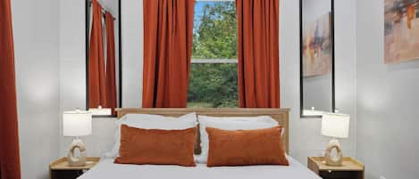 A hotel room with a bed, two pillows, a window with curtains, a bedside table with a lamp, and a mirror on the wall.