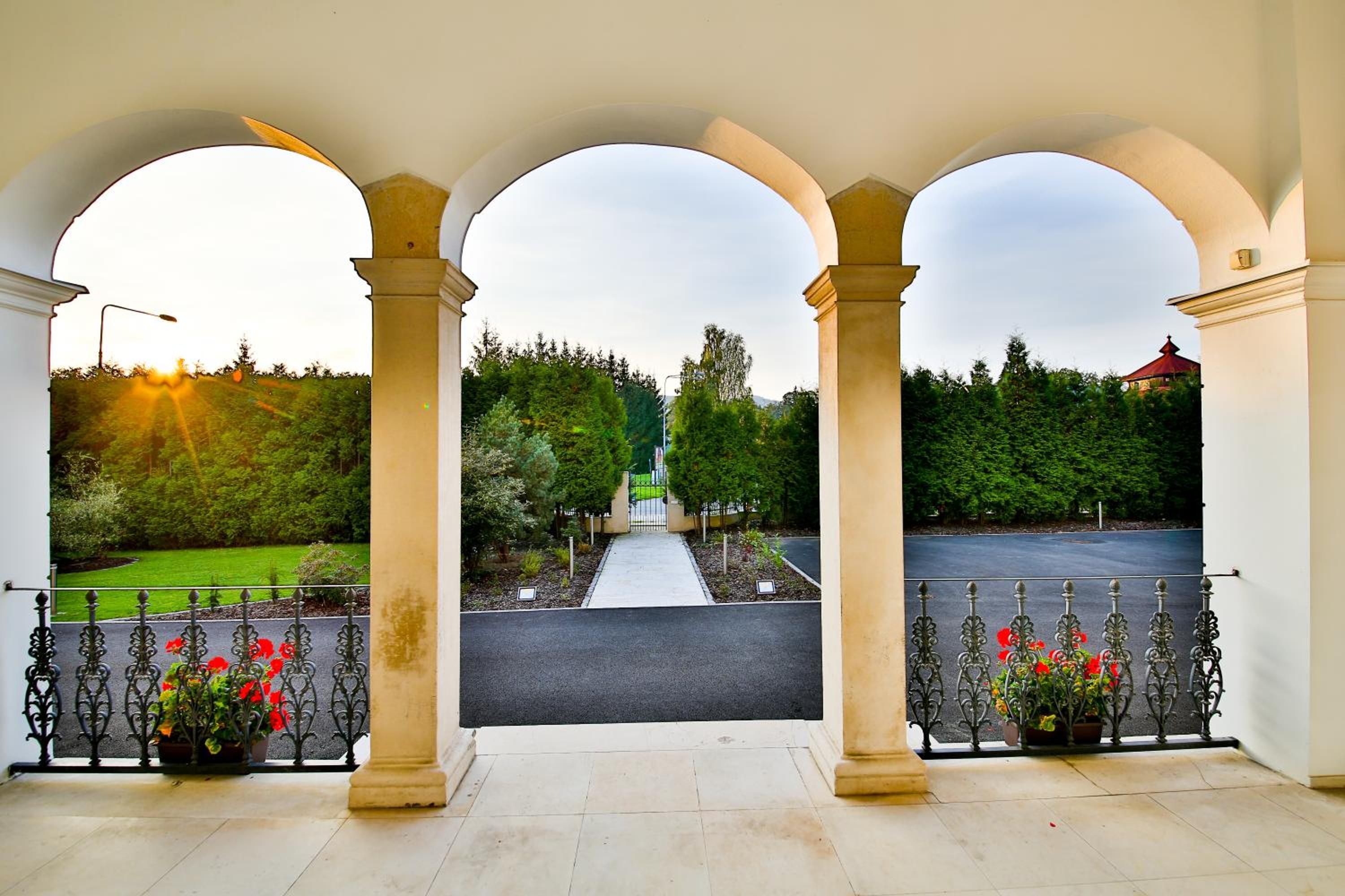 Una entrada cubierta con tres arcos, un camino que lleva a una puerta y una vista de paisaje verde con árboles y un edificio en la distancia.