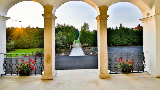Una entrada cubierta con tres arcos, un camino que lleva a una puerta y una vista de paisaje verde con árboles y un edificio en la distancia.