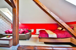A bedroom with a sloped ceiling, a bed with red bedding, a grey sofa with red cushions, and a white radiator.