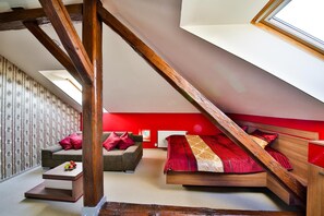 A loft bedroom with a sloped ceiling, a wooden staircase, a bed with red bedding, a grey sofa, and a small table with fruit.