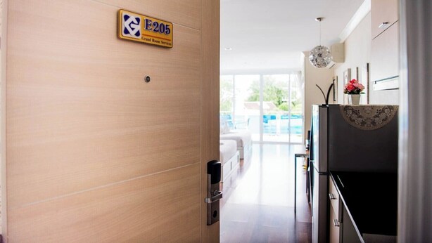Quarto de hotel com porta de madeira, placa indicando o número do quarto E205, uma cama, uma TV e vista para o exterior.