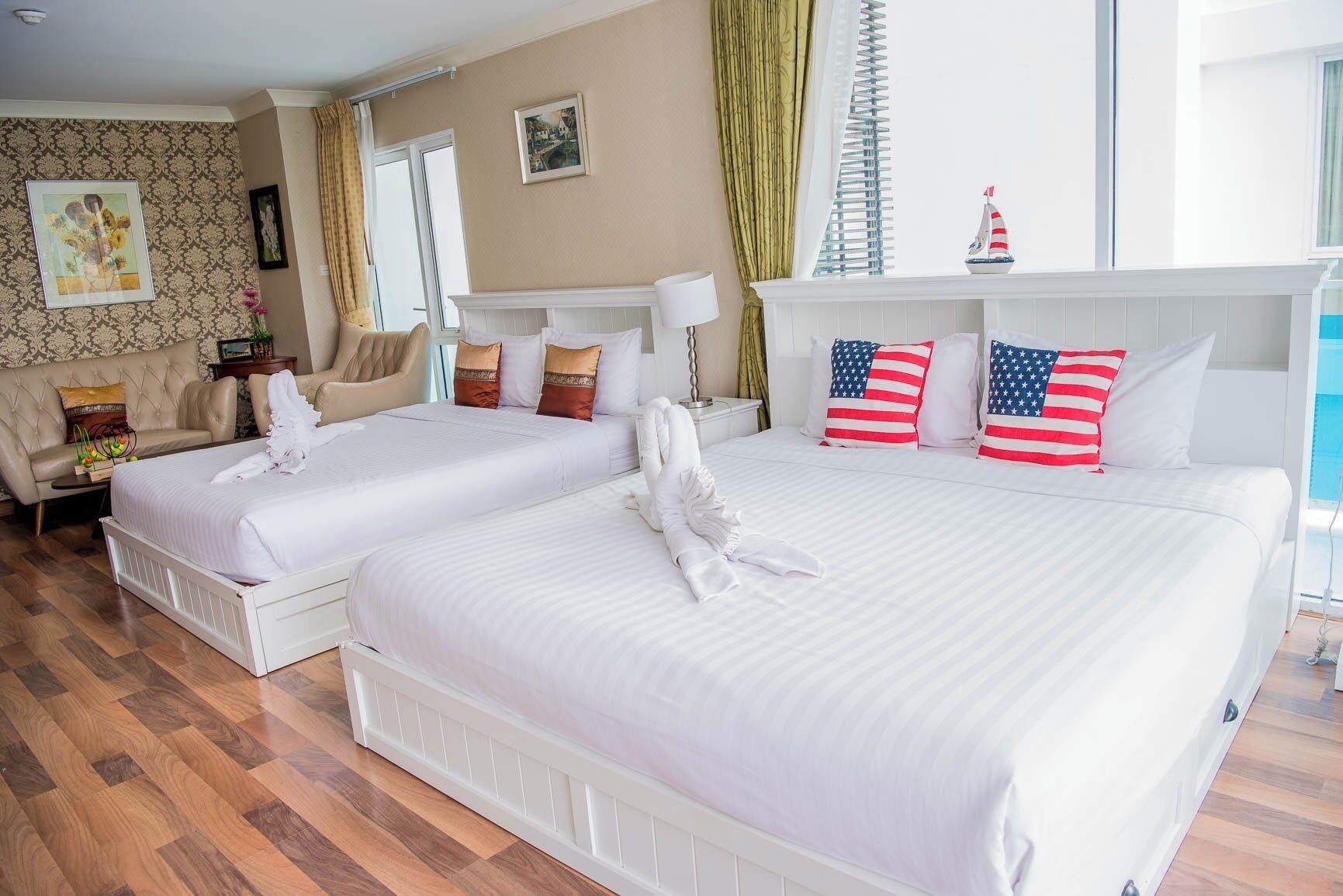 A hotel room with two beds, a sofa, a window with curtains, and a small sailboat decoration.