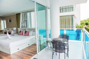 Habitación de hotel con cama, una mesita, dos sillas, balcón con cerramiento de vidrio con piscina y vista a zonas verdes.