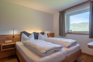 Una cama doble con ropa blanca, cabecera de madera, lámparas de noche y una ventana con vistas a un bosque.