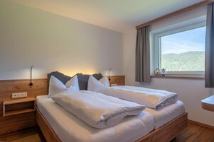 Una cama doble con ropa blanca, cabecera de madera, lámparas de noche y una ventana con vistas a un bosque.