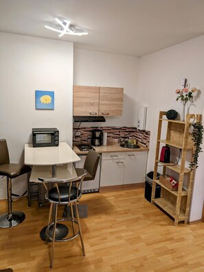 Een compacte keuken met een magnetron, broodrooster en koffiezetapparaat. Er is een eettafel met stoelen en een klein krukje. Een houten wandplank is tegen de muur geplaatst.