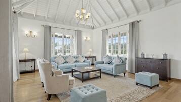 A spacious living room with a high ceiling, a chandelier, a sofa set, a coffee table, a side table, a lamp, a cabinet, and a window with curtains.