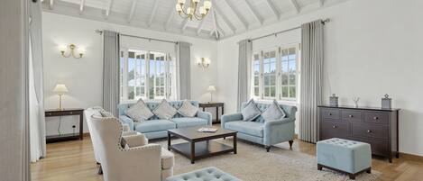 A spacious living room with a high ceiling, a chandelier, a sofa set, a coffee table, a side table, a lamp, a cabinet, and a window with curtains.