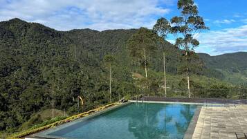 A rectangular pool with stone steps leading up to it, surrounded by a stone wall, set against a backdrop of lush green hills and a clear blue sky.