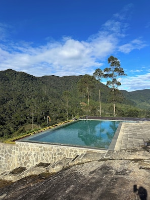 A rectangular pool with stone steps leading up to it, surrounded by a stone wall, set against a backdrop of lush green hills and a clear blue sky. - Aadya Tea Bangalows By Aahaasa (Upcot)
