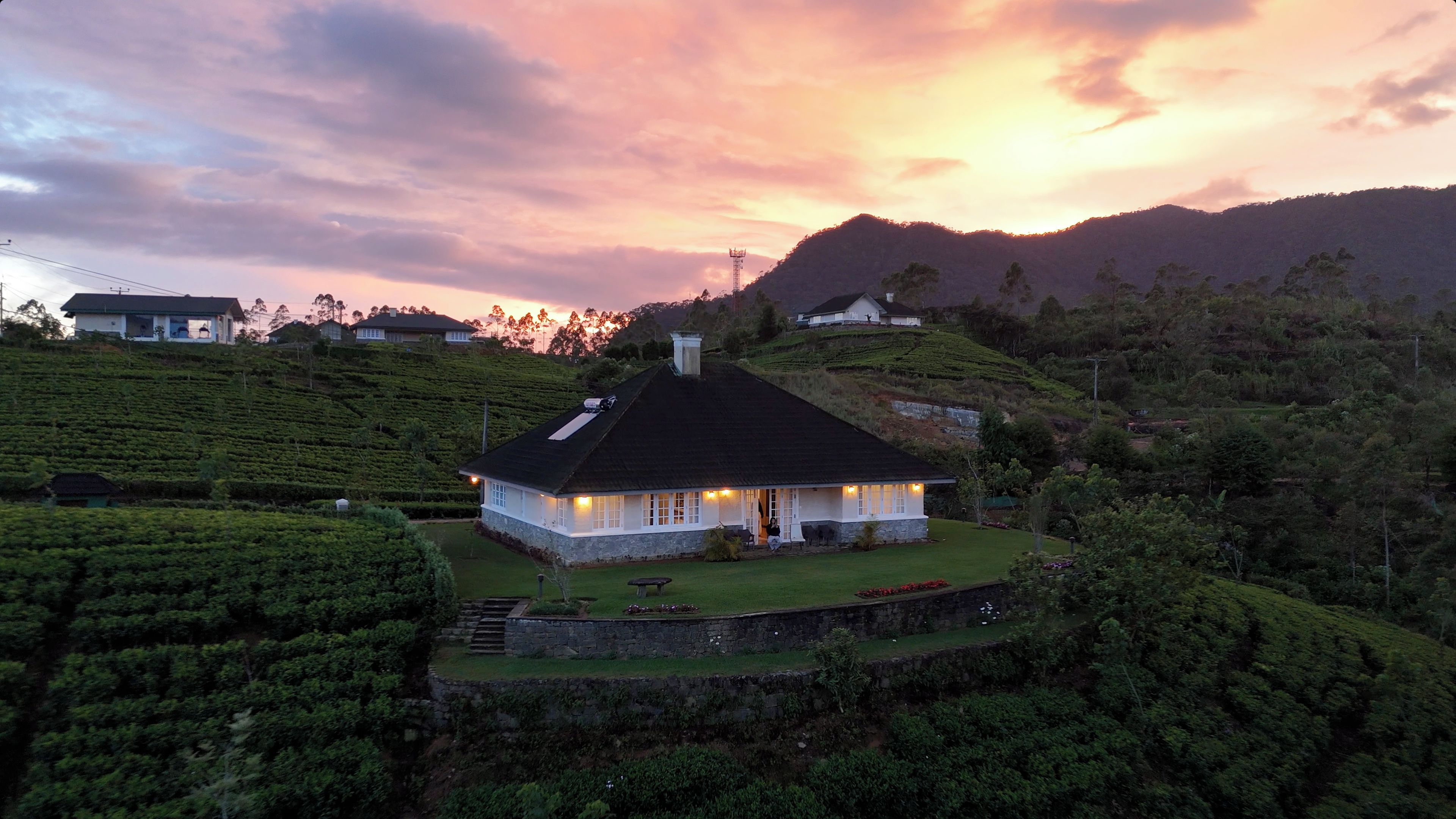 A house with a stone foundation, surrounded by lush green tea plantations, set against a backdrop of hills and a sunset sky.