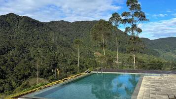 A rectangular pool with stone steps leading up to it, surrounded by a stone wall, set against a backdrop of lush green hills and a clear blue sky.