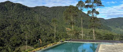 A rectangular pool with stone steps leading up to it, surrounded by a stone wall, set against a backdrop of lush green hills and a clear blue sky.