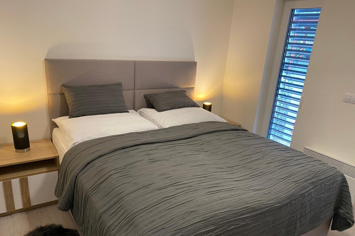 Un dormitorio con cama, dos lámparas de noche, una ventana con persianas y una alfombra pequeña.