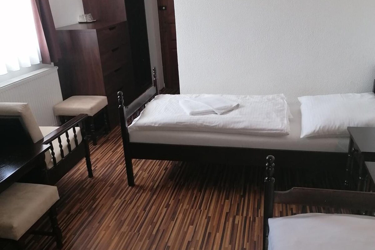 Una habitación de hotel con piso de madera, una cama con ropa blanca, un banco de madera y una lámpara de pared.