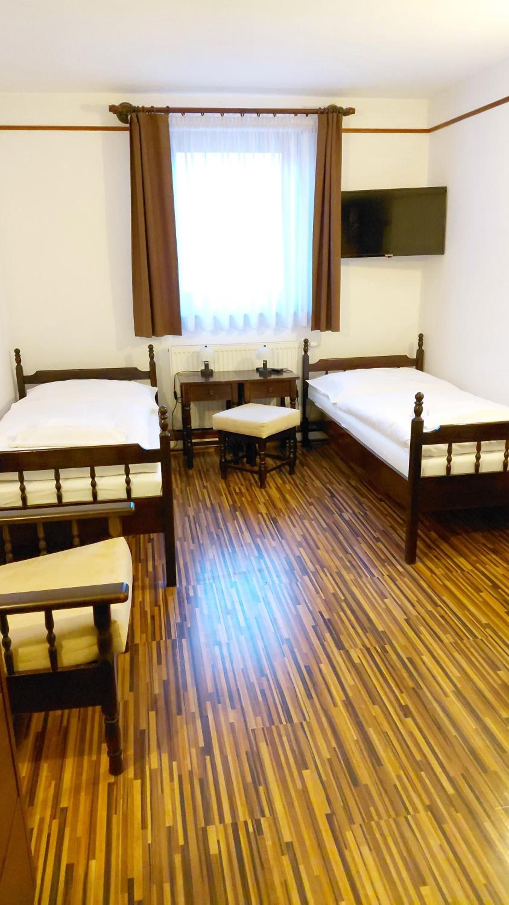 Habitación de hotel con dos camas individuales, suelo de madera, un escritorio con silla, una ventana con cortinas y un televisor de pantalla plana montado en la pared.