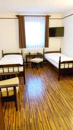 Habitación de hotel con dos camas individuales, suelo de madera, un escritorio con silla, una ventana con cortinas y un televisor de pantalla plana montado en la pared.