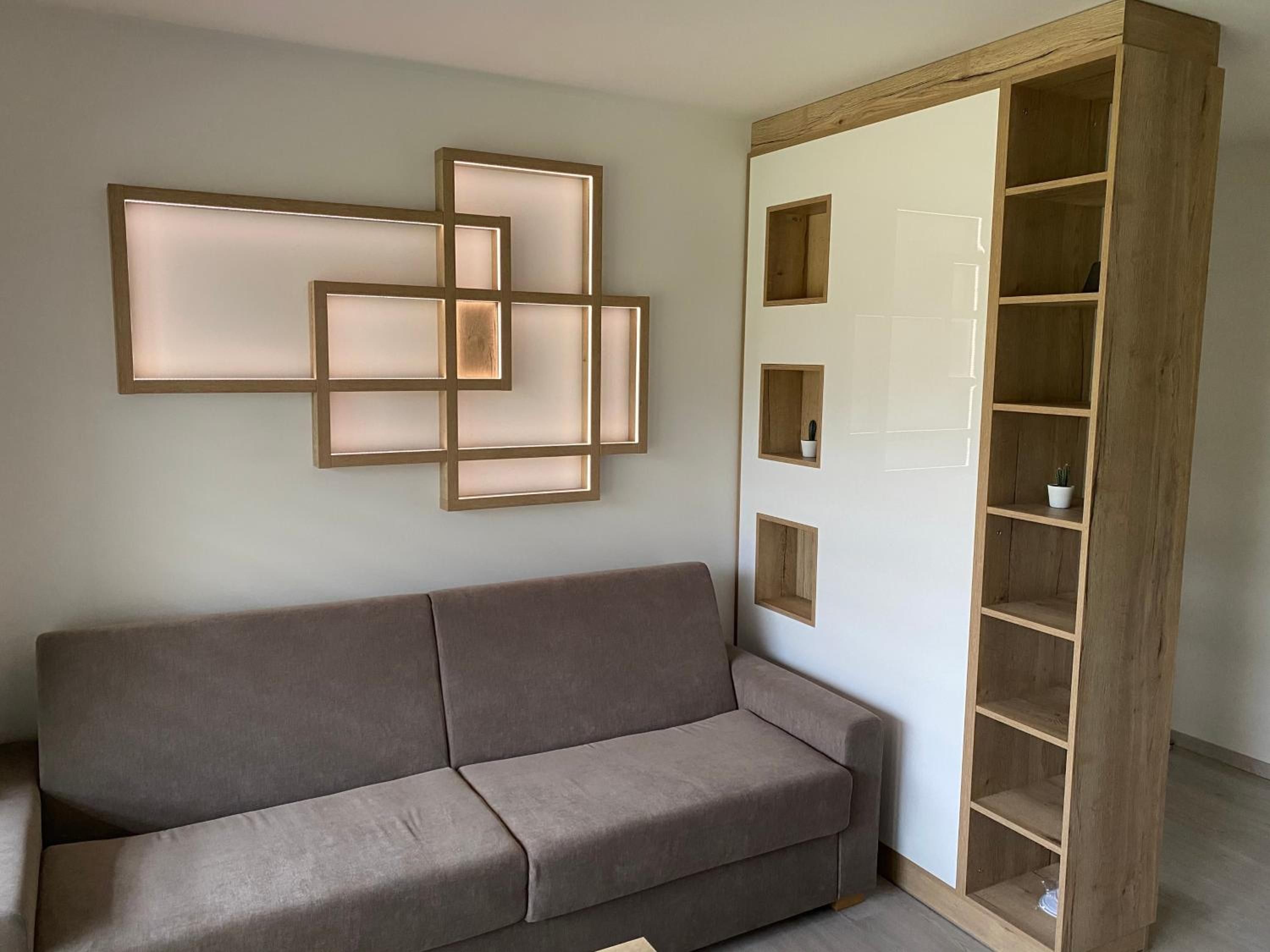 Una habitación en tonos neutros con un sofá gris, una estantería de madera con varios compartimentos y una lámpara fijada a la pared.