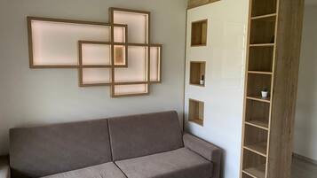 Una habitación en tonos neutros con un sofá gris, una estantería de madera con varios compartimentos y una lámpara fijada a la pared.