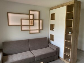 Una habitación en tonos neutros con un sofá gris, una estantería de madera con varios compartimentos y una lámpara fijada a la pared.
