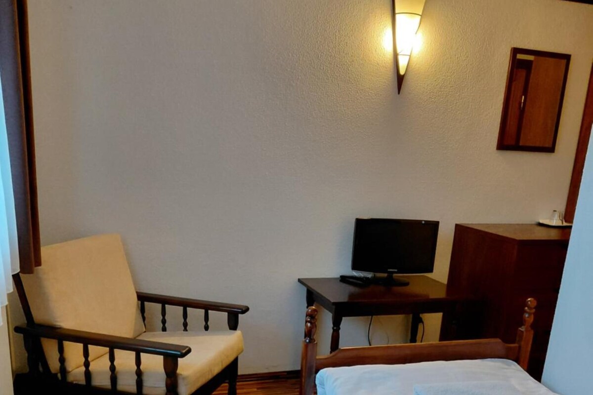 A hotel room with a wooden floor, a bed with white linens, a chair, a desk with a television, and a wall-mounted light fixture.