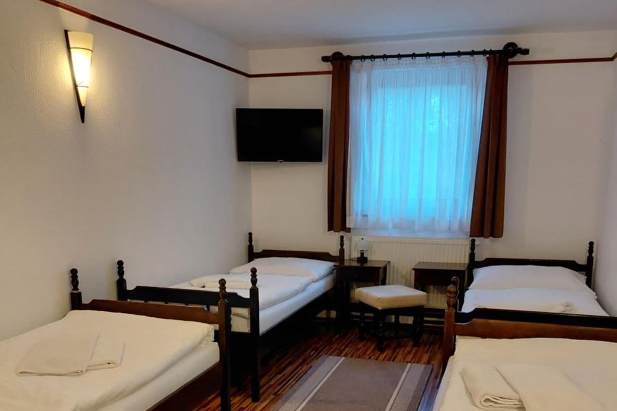 A hotel room with two single beds, a flat-screen TV mounted on the wall, a window with curtains, and a wooden floor.